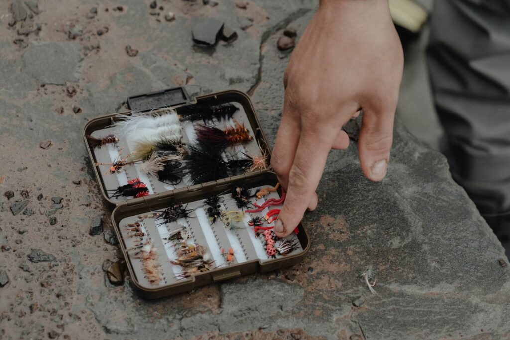 A small box of fishing lures sits on a slate wall. It contains multiple rows of colorful and furry lures. A person points to one of the bottom rows, touching the lures.