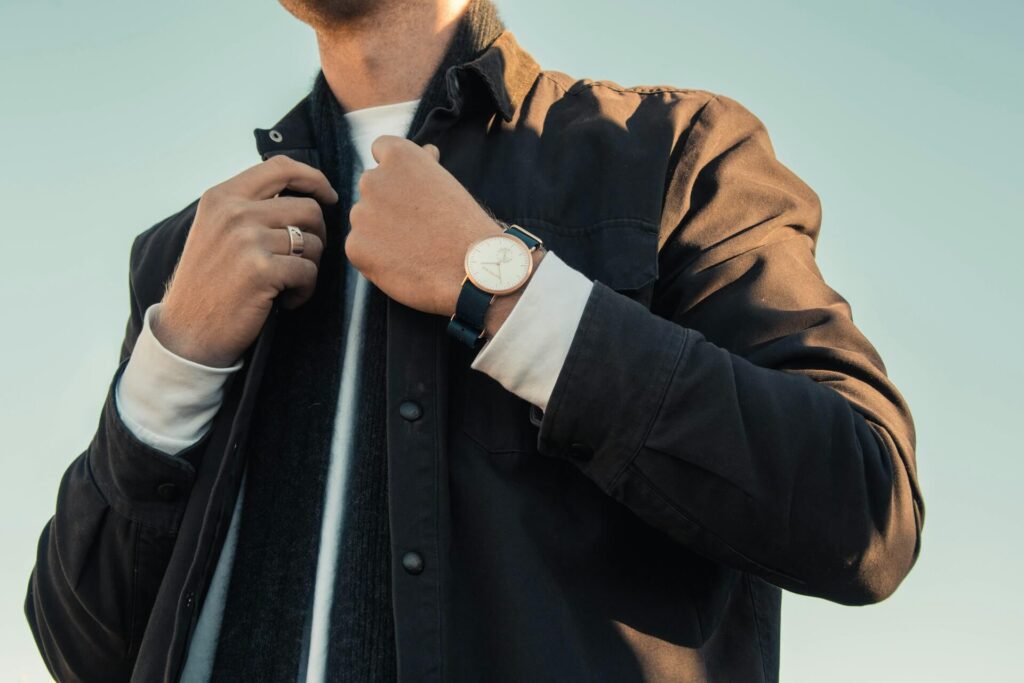 A man stands holding the two middle seams of his brown denim jacket. He wears one gold ring, a gold watch on a black band, a white dress shirt, and a gray scarf underneath his jacket.