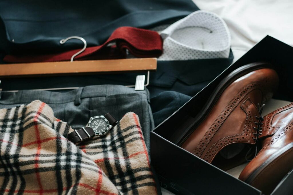 A collection of men's clothes sits on a bed. On top of the white duvet is gray slacks on a pants hanger, a black suit jacket over a polka dotted white button down, a red tie and a black shoe box with brown loafers. On top of the gray slacks is a brown, red, white and black checkered scarf and a watch on a brown band.