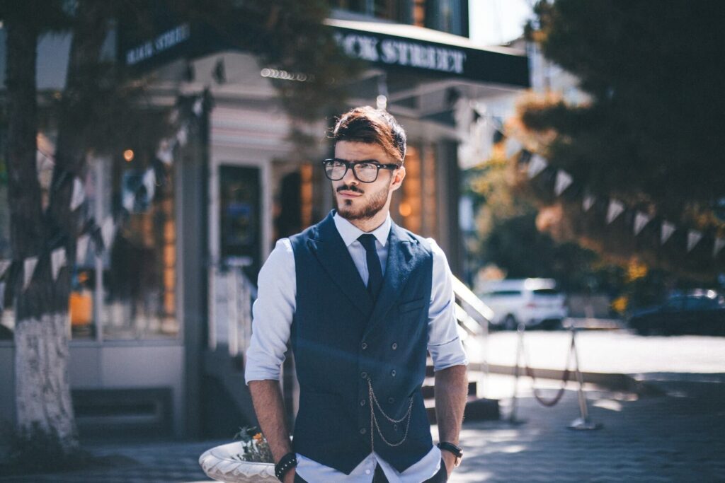 A man stands in front of a business on a city street. He peers into the distance while wearing black rimmed glasses, a white dress shirt with rolled up sleeves, a black skinny tie, a navy vest, a gold chain attacked to the vest's buttons, a black wrist band and a gold watch on a black leather band.