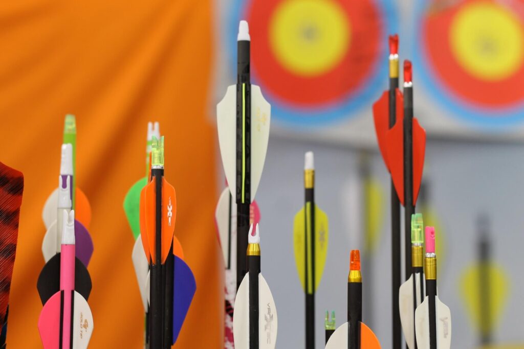 A close-up of arrow fletchings. Most of the arrows have black stems, while the fletchings are all different colors. Out-of-focus targets are in the background.