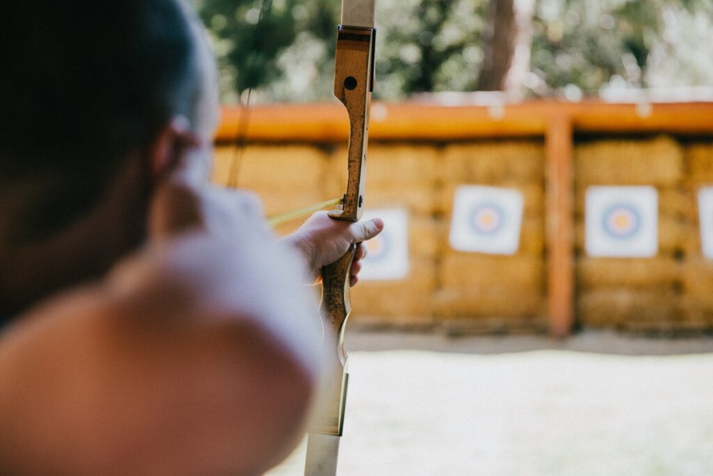 An over-the-shoulder profile of a man with a bow drawn tight. He aims at a target hanging in front of a wooden wall, surrounded by other hanging targets.