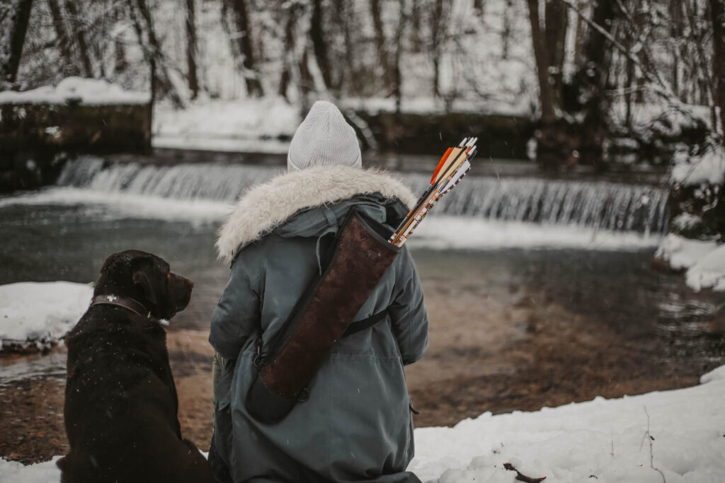 A person in a winter coat and white knitted cap sits facing a rushing river with a small waterfall cutting through the middle. The person wears a leather quiver with a few arrows inside it. A winter forest background surrounds the river while a chocolate lab with a leather collar sits next to the archer at rest.