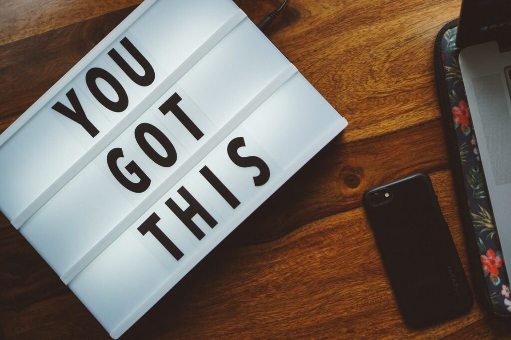 A crooked white sign with black text reading You Got This sits on a wooden table. The corner of a laptop is seen on a laptop pad featuring a floral print. A phone in a black case lies face down on the wooden table.