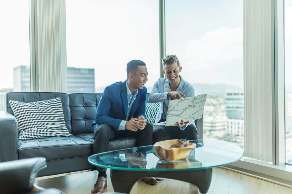 Two men sit on a leather couch in front of floor-to-ceiling windows in the corner of an apartment high in a sky riser. An unknown city landscape is behind the. The man on the right points to the screen of the laptop he's holding so the man on the left can see. They're both laughing.