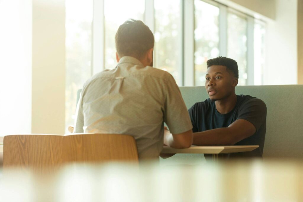 Two individuals sit at a table engaged in conversation with a bright window in the background. The person facing the camera is wearing a blue t-shirt and seated on a couch, while the other person has their back to the camera and is wearing a light gray shirt.