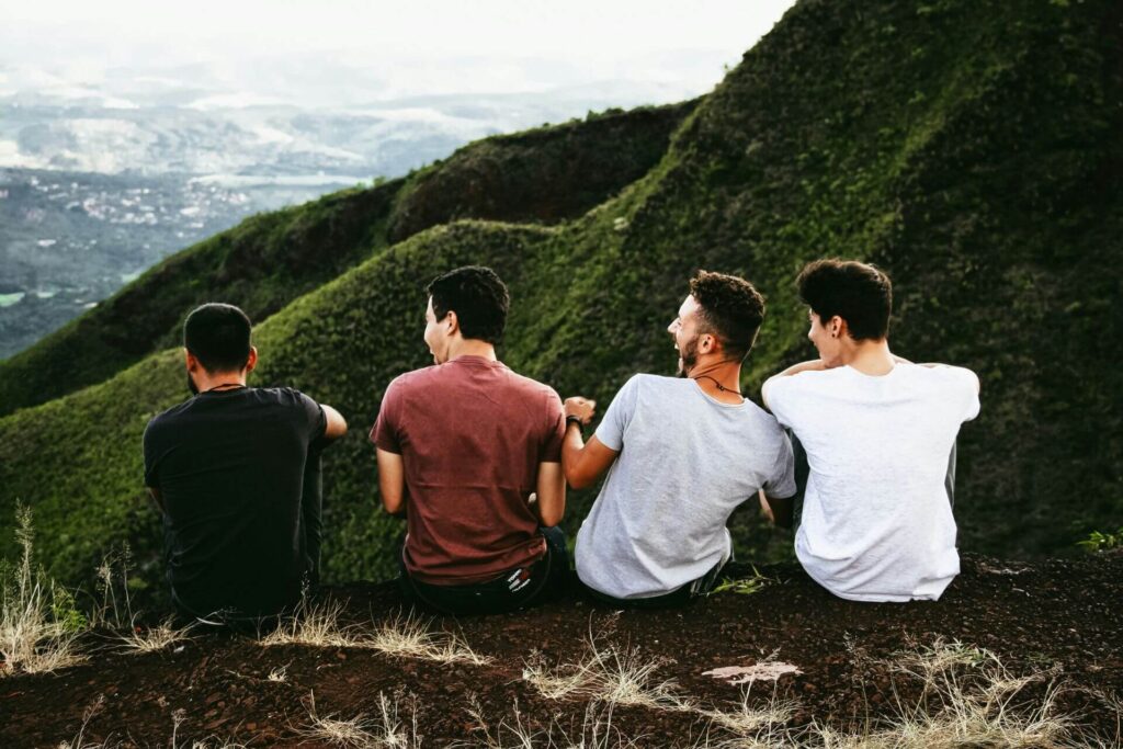 Three people sitting closely together on a green mountain slope, overlooking a panoramic view of a valley with patches of residential areas and winding roads. The person on the left is wearing a black shirt, the middle one is in a maroon shirt, and the one on the right is in a white shirt. They appear to be in a relaxed and contemplative mood, enjoying the vast landscape stretching before them.