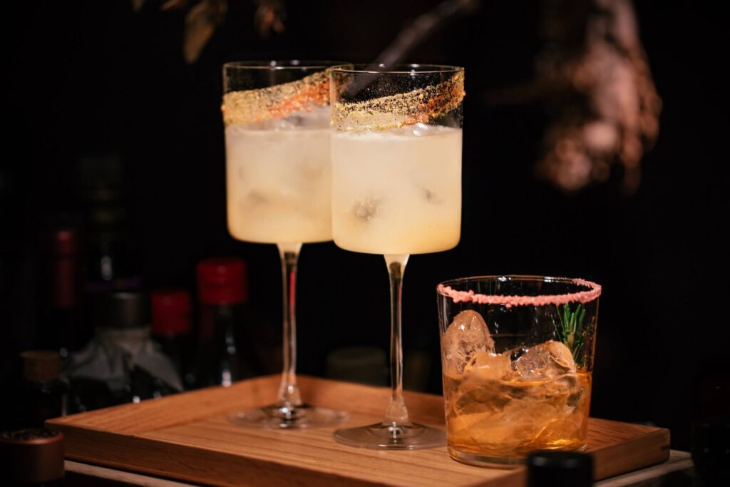 Two tall cocktail glasses stand on a wooden board. They contain a yellow beverage with ice, plus a cinnamon sugar rim around the glass. A smaller whiskey glass filled with caramel colored liquid and ice sits next to it. It has a pink salt rim and a green garnish sprig inside it.