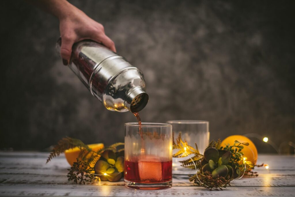 A person pours a red cocktail solution out of a stainless steel shaker into a short glass with a large square ice cube. Greenery and fairy lights surrounds the glass. Lemons and an empty glass sit in the background.