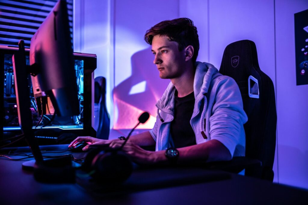 A guy plays on his PC while sitting in a gaming chair in a room lit with purple and blue lighting.