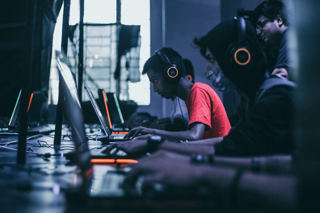 A guy plays games on a laptop surrounding by other people doing the same thing. He wears a red shirt and orange headphones. People watch behind them.